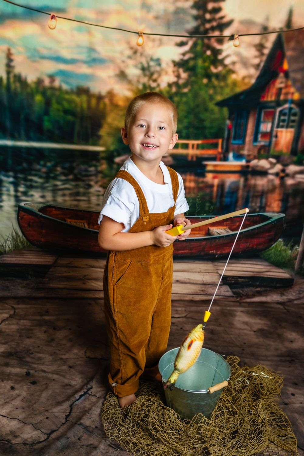 Child in brown overalls sits on a wooden dock by a rustic lake house with a boat at sunset.