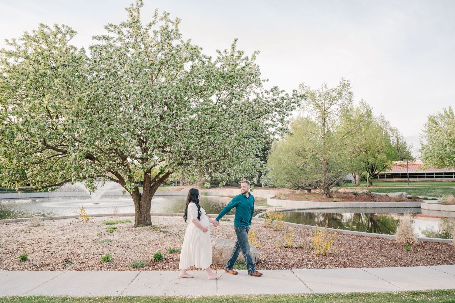 A couple in formal attire holds hands while walking through a blooming orchard garden in springtime.