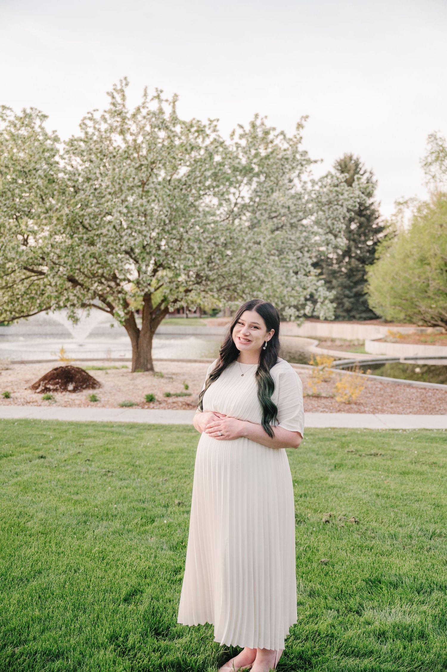 Person in a white pleated maxi dress stands outdoors on green grass with a flowering tree in the background.