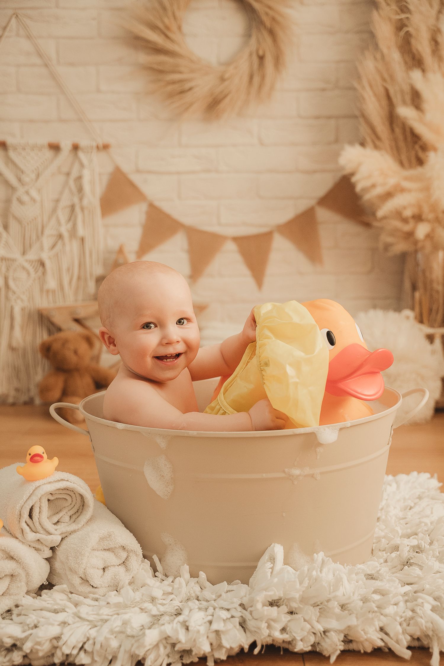 Smiling baby sits in miniature white bathtub with yellow rubber duck toy surrounded by rustic farmhouse decor.