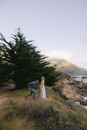 Two people share a romantic moment on a scenic coastal cliff with evergreen trees and misty mountains in the background.