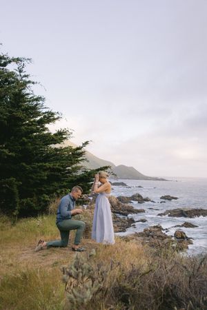 A romantic proposal scene unfolds on a cliff overlooking the rugged California coastline with crashing waves below.
