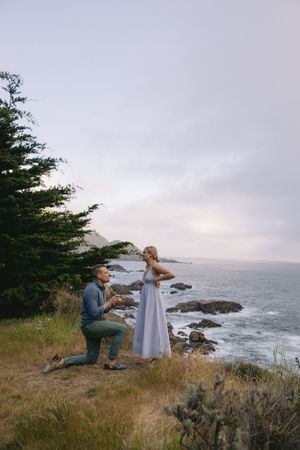 A couple shares a romantic proposal moment on a coastal cliff overlooking the ocean at sunset.