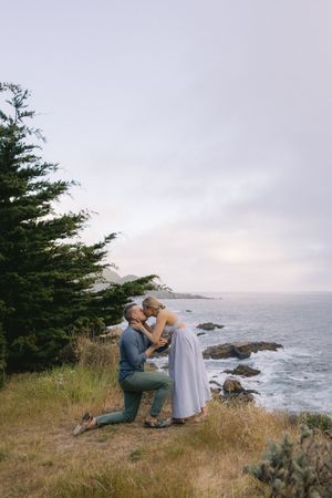 A romantic engagement moment on a scenic coastal cliff overlooking the Pacific Ocean with evergreen trees and rocky shores.