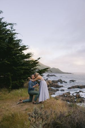 Two people share an intimate moment on a cliff overlooking a rocky Pacific coastline at dusk.