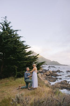 A proposal moment unfolds on dramatic California coastline with cypress trees and rocky shores at dusk.