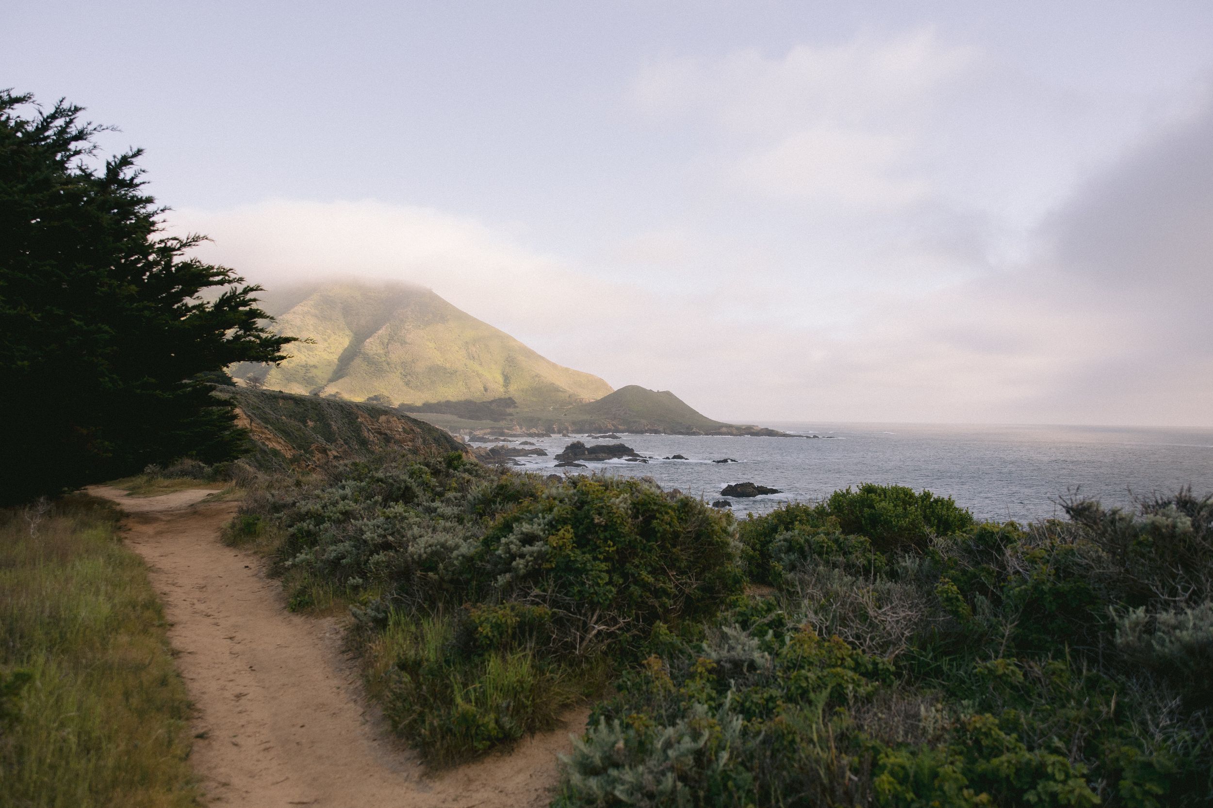 A dirt hiking trail winds along the rocky California coastline with mountain views in the misty distance.