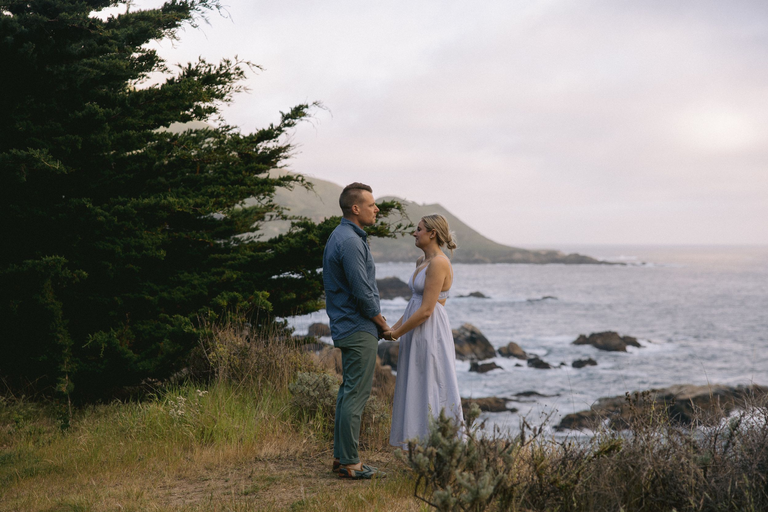 A romantic couple stands on a coastal cliff overlooking the Pacific Ocean with rocky shores and mountains in the distance.
