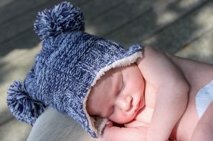 Sleeping baby wearing a cozy knitted blue winter hat with pom poms and white trim on soft blanket.