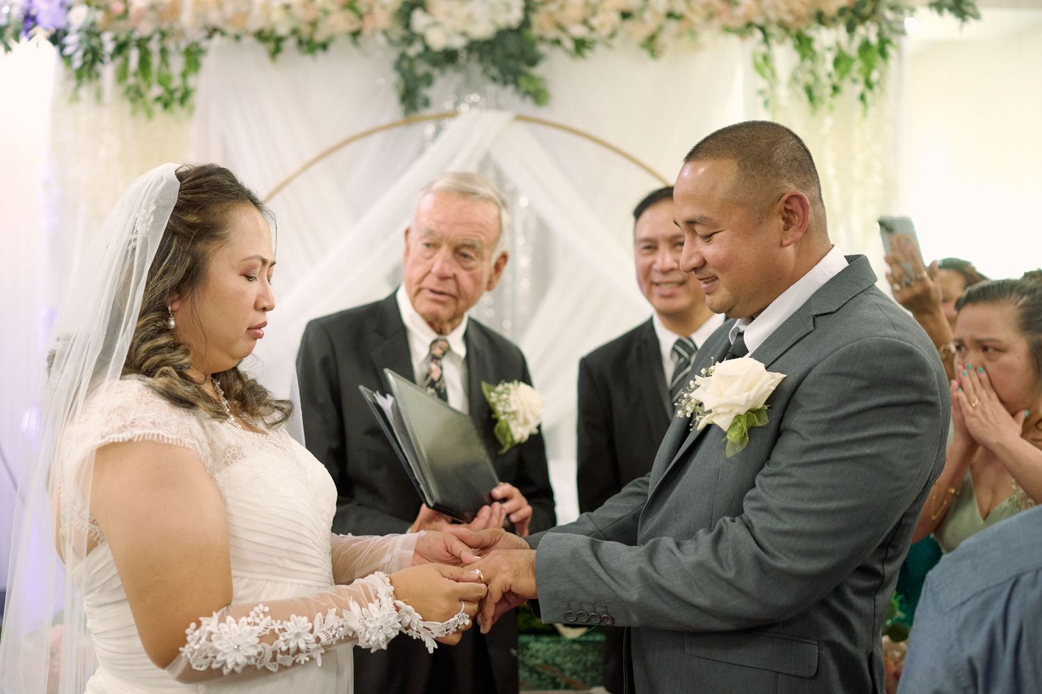 A couple exchanges rings during a wedding ceremony while an officiant presides under floral decorations.