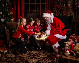 Santa Claus shows children a golden glowing gift box in a cozy Christmas-themed room with red decor and presents.