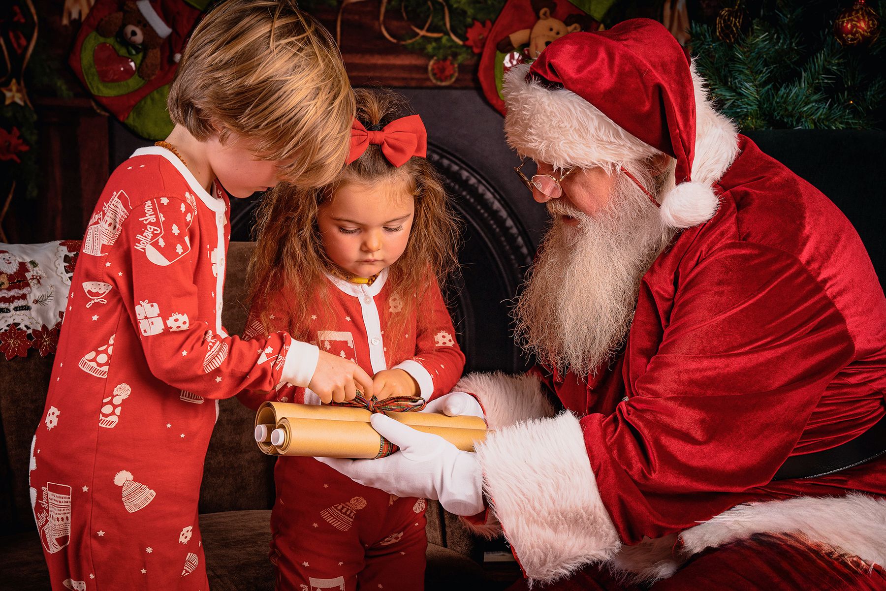 Children in matching red pajamas look at a book with Santa Claus during holiday photo session.