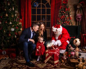 A cozy Christmas scene with Santa Claus showing a present to a small child by decorated trees and red curtains.
