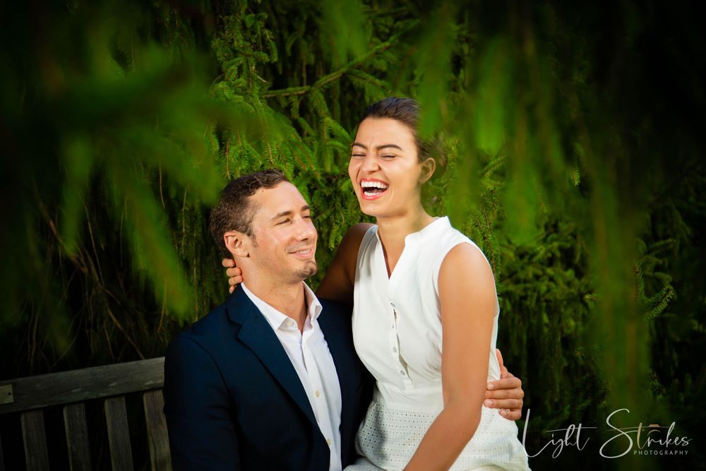 A joyful couple embraces and laughs together in front of lush green foliage during an outdoor photo session.