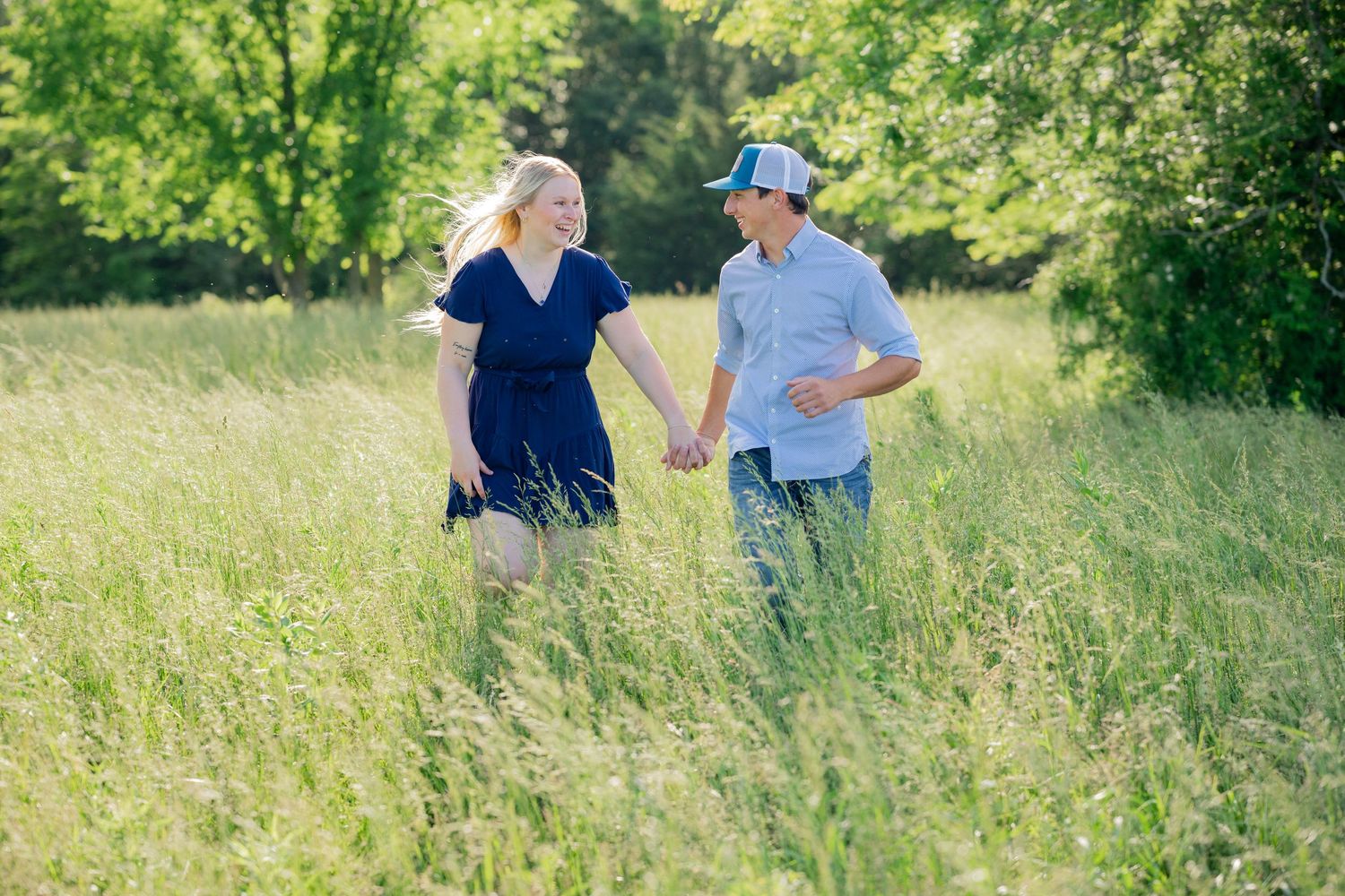Couple walking hand in hand through a sunlit meadow with tall grass.