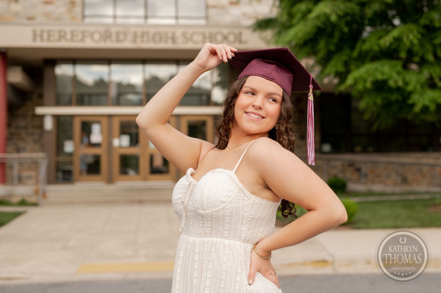 Graduate in maroon cap playfully poses in white dress near school entrance.
