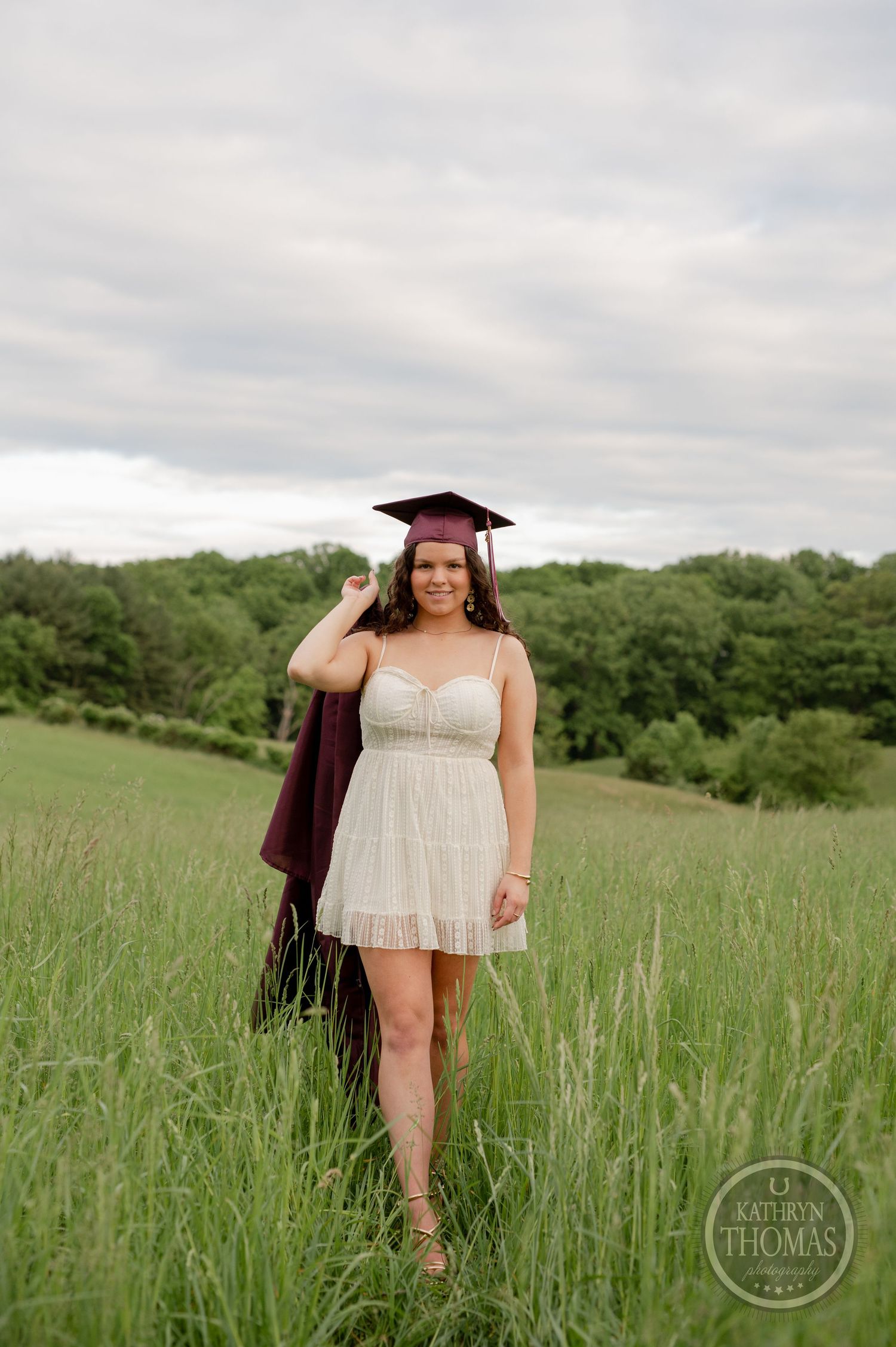 Graduate walks through tall grass wearing white dress and maroon graduation attire.