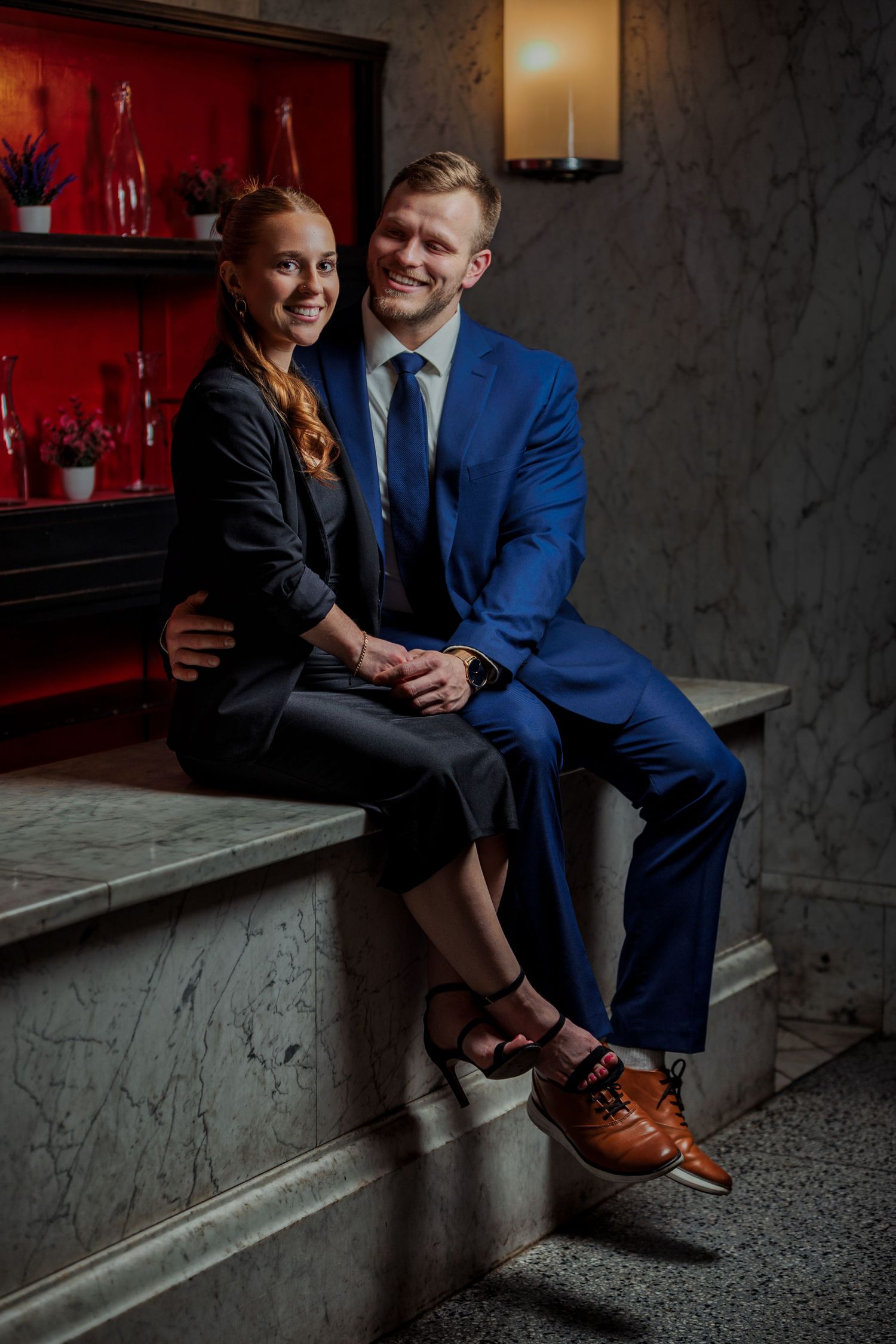 Smiling couple in formal attire sitting together on marble steps in an elegant interior setting.