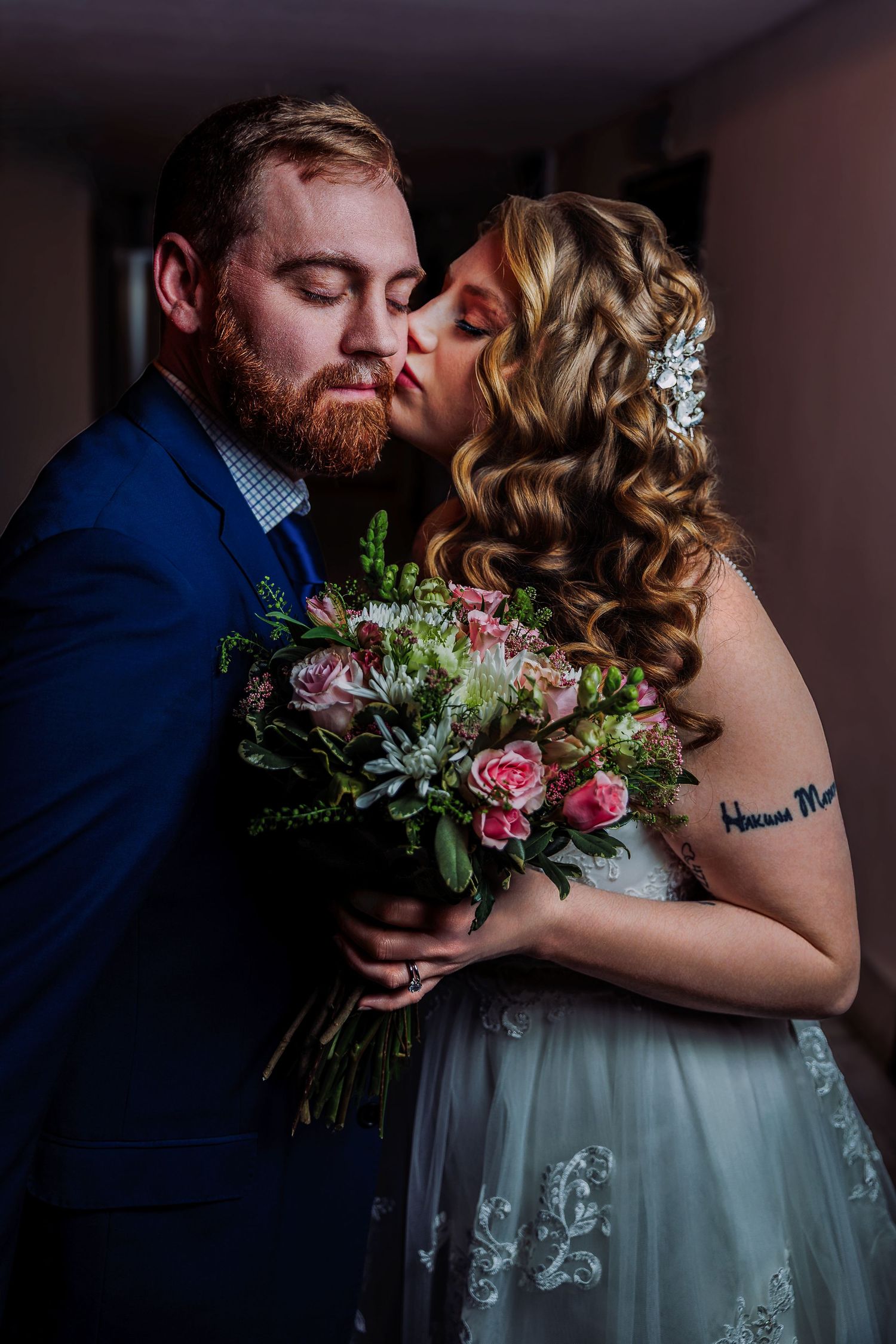Bride kisses groom on cheek as they hold a pink rose bouquet, both dressed in wedding attire in a romantic portrait.