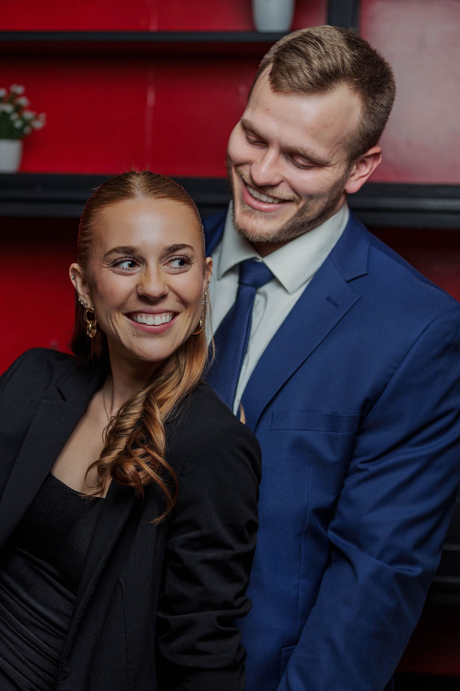A smiling couple in formal attire, woman in black dress and man in blue suit, posing together against a red background.