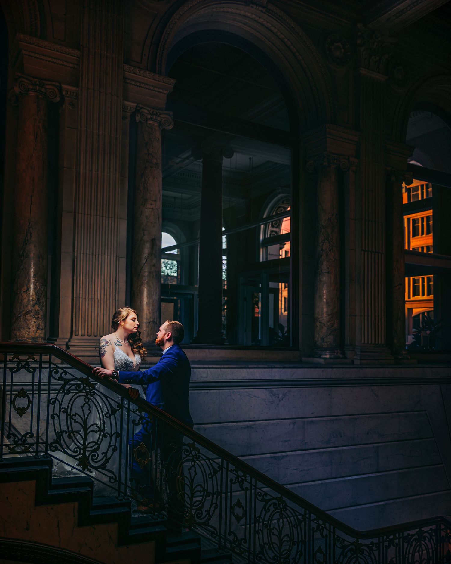 A couple in formal attire shares a romantic moment on an ornate staircase in a grand historic building.
