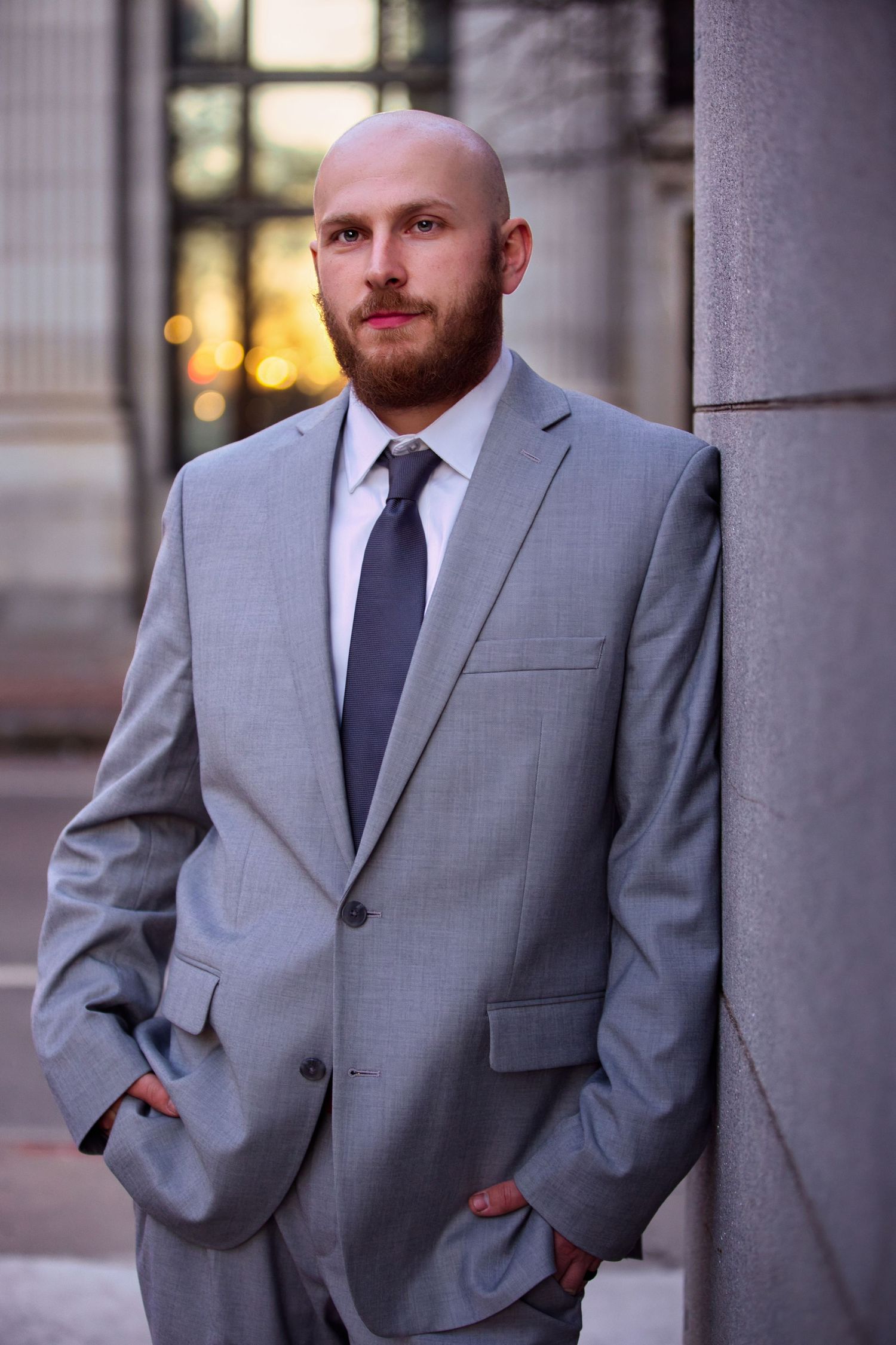 Bald bearded man in gray suit and tie leaning against column outdoors with bokeh city lights background.