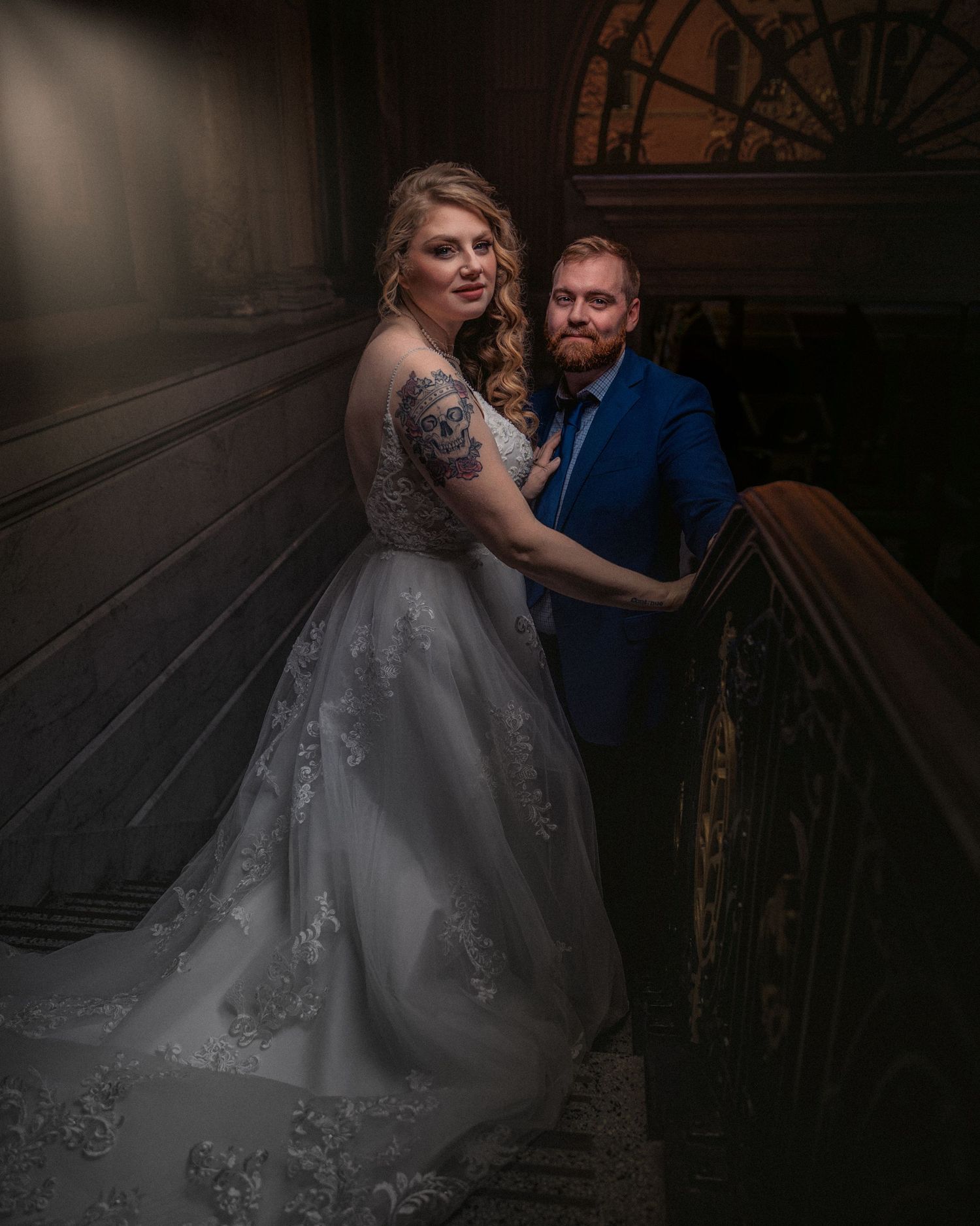 Bride in white lace gown and groom in blue suit pose elegantly on grand staircase in ornate venue.