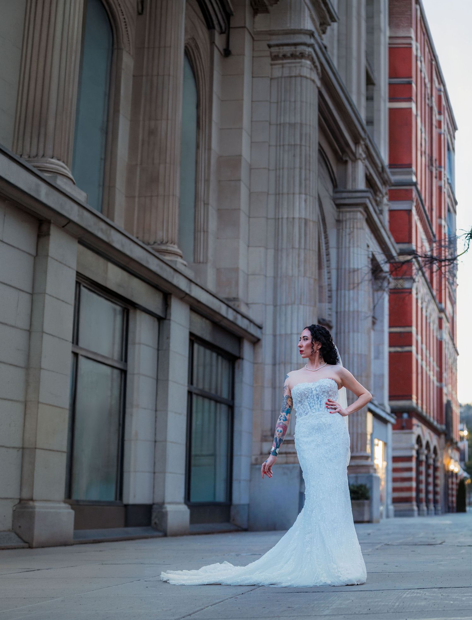 Bride in white mermaid gown poses on urban sidewalk beside grand stone building at dusk.