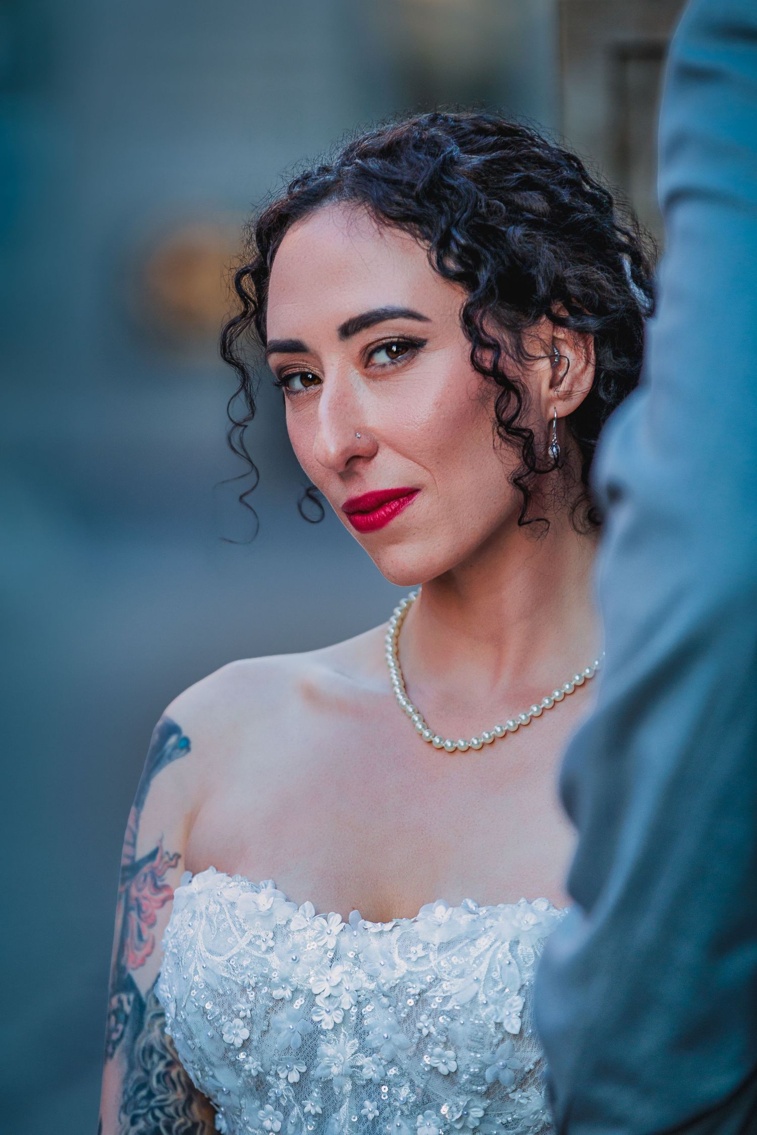 Bride with curly hair, red lips, pearl necklace, and floral strapless gown with tattoo visible on shoulder.