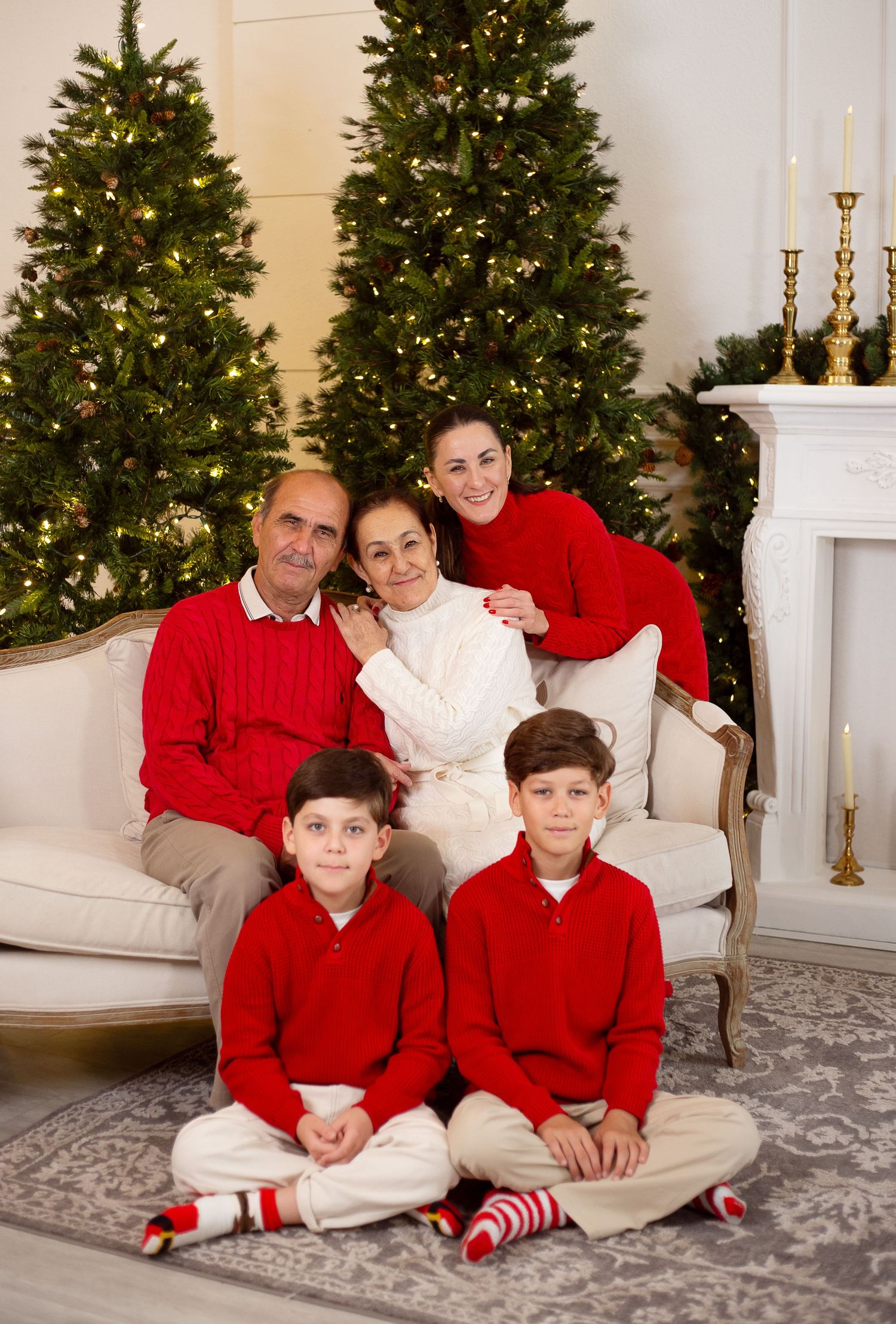 A festive holiday family portrait in front of two Christmas trees and a fireplace, all wearing red and white outfits.