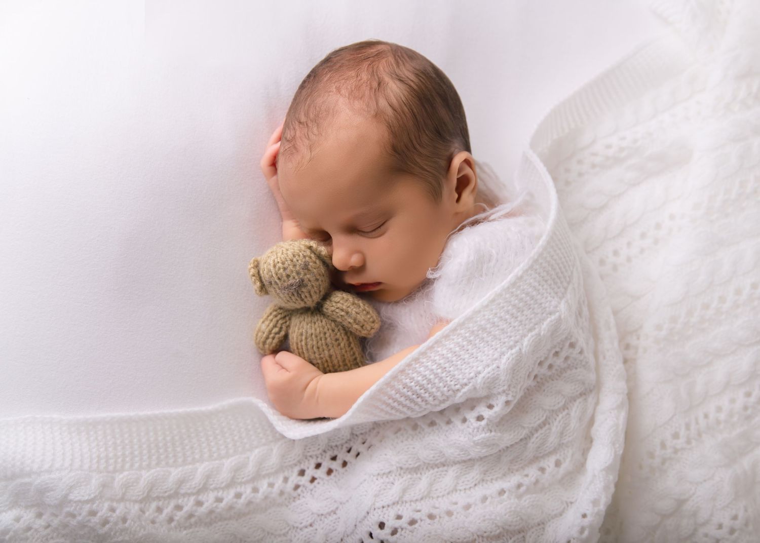 Newborn asleep under blanket cuddling brown teddy bear.
