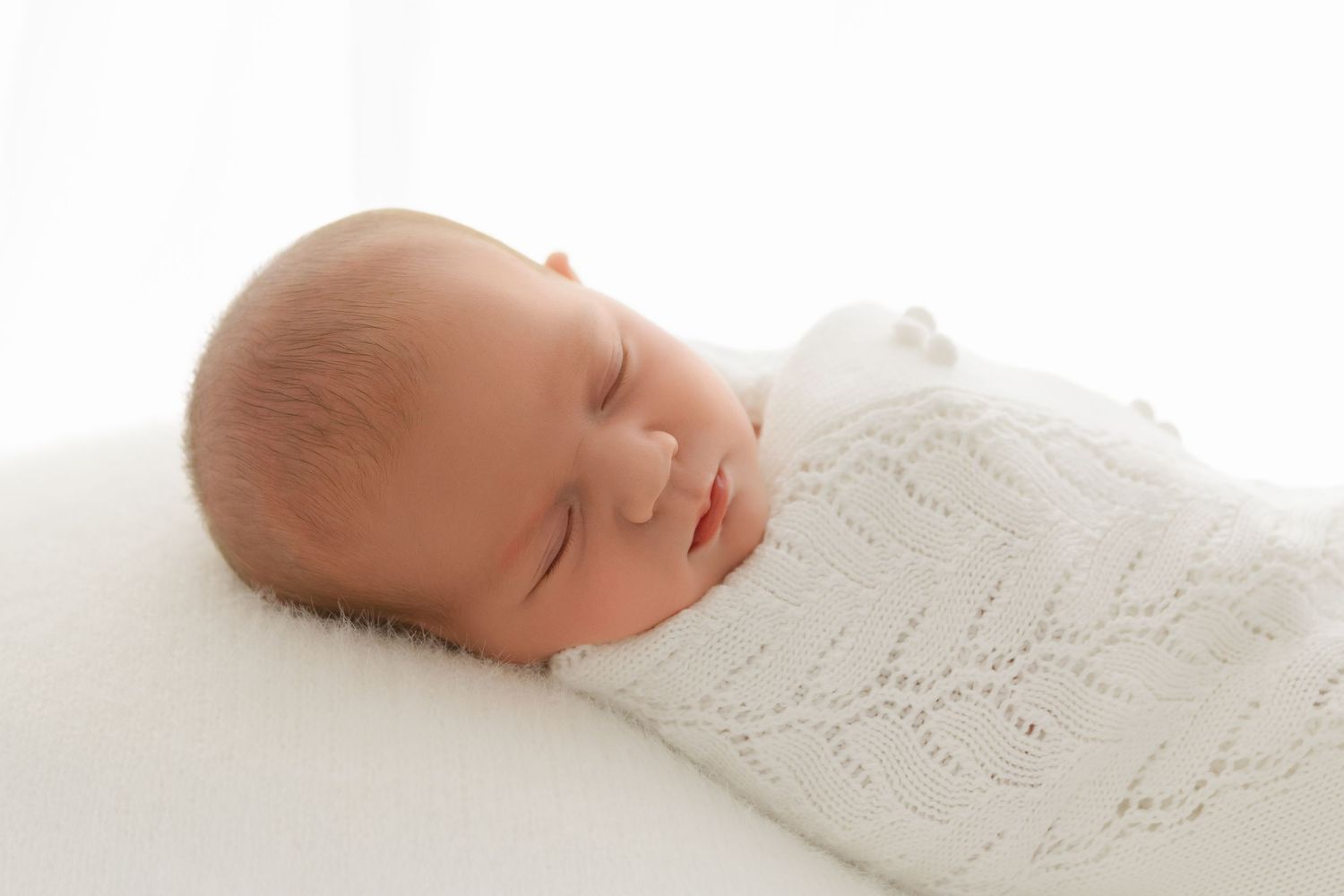 Peaceful newborn baby swaddled in white knit wrap sleeping on soft white pillow.