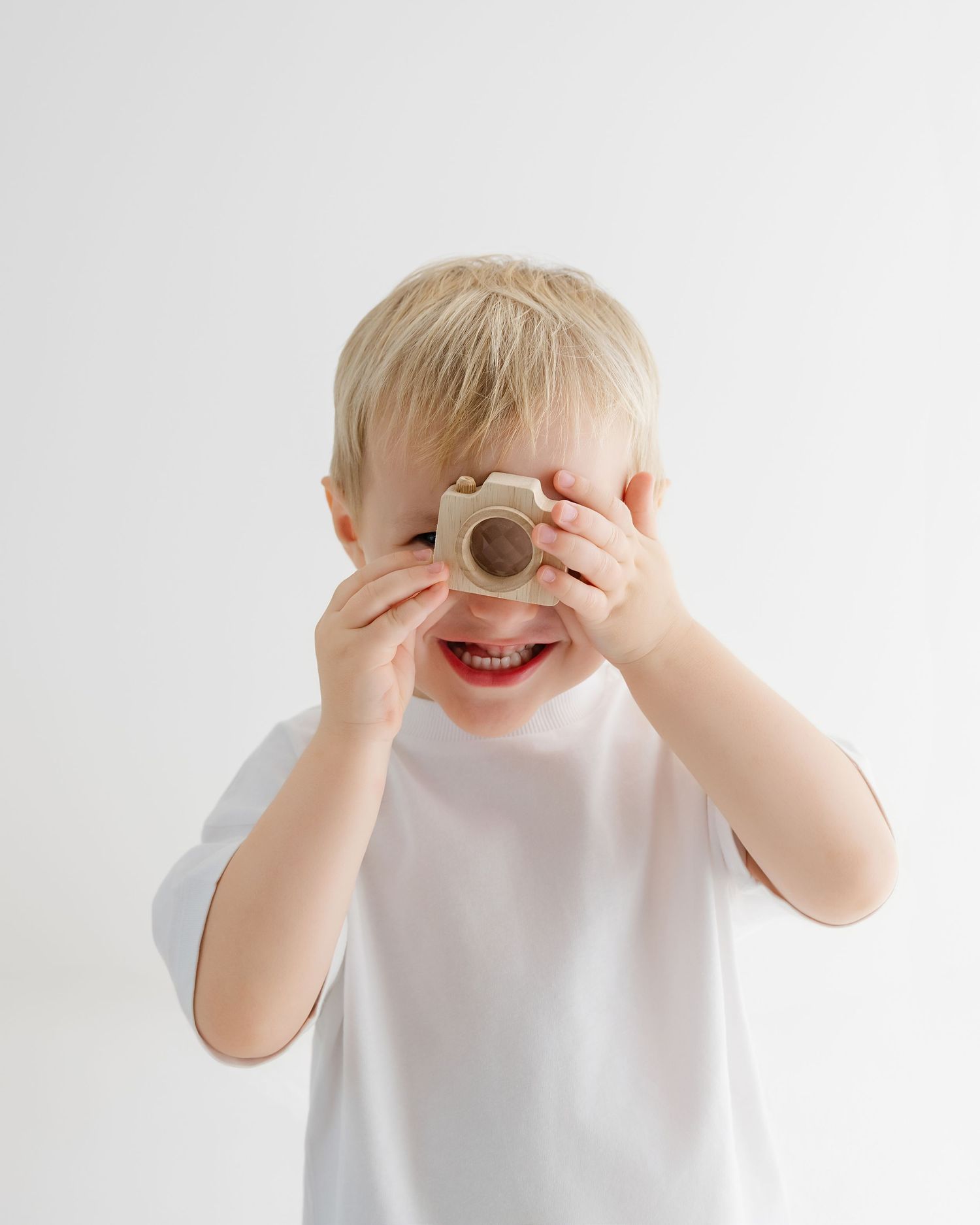 Smiling young boy in white t-shirt holding a small wooden toy camera up to his face against white background.
