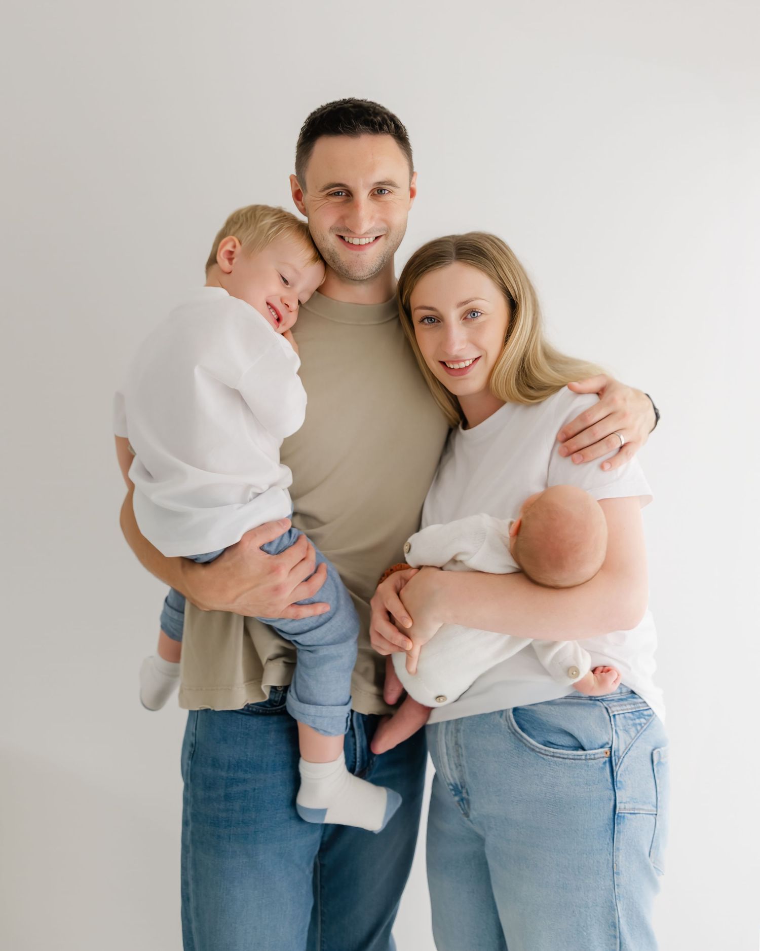 A smiling family of four poses together, parents holding a toddler and newborn baby, dressed in white and denim.
