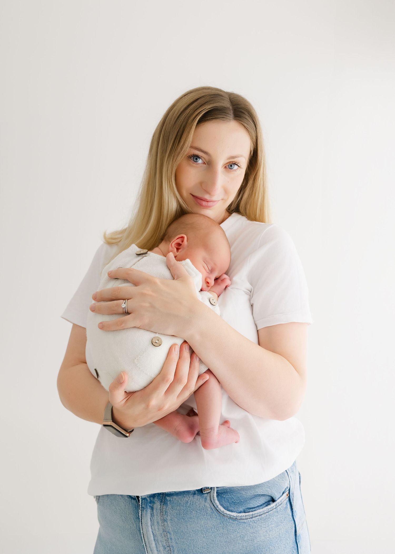 A mother with blonde hair holds her sleeping newborn baby close to her chest in a bright white studio setting.