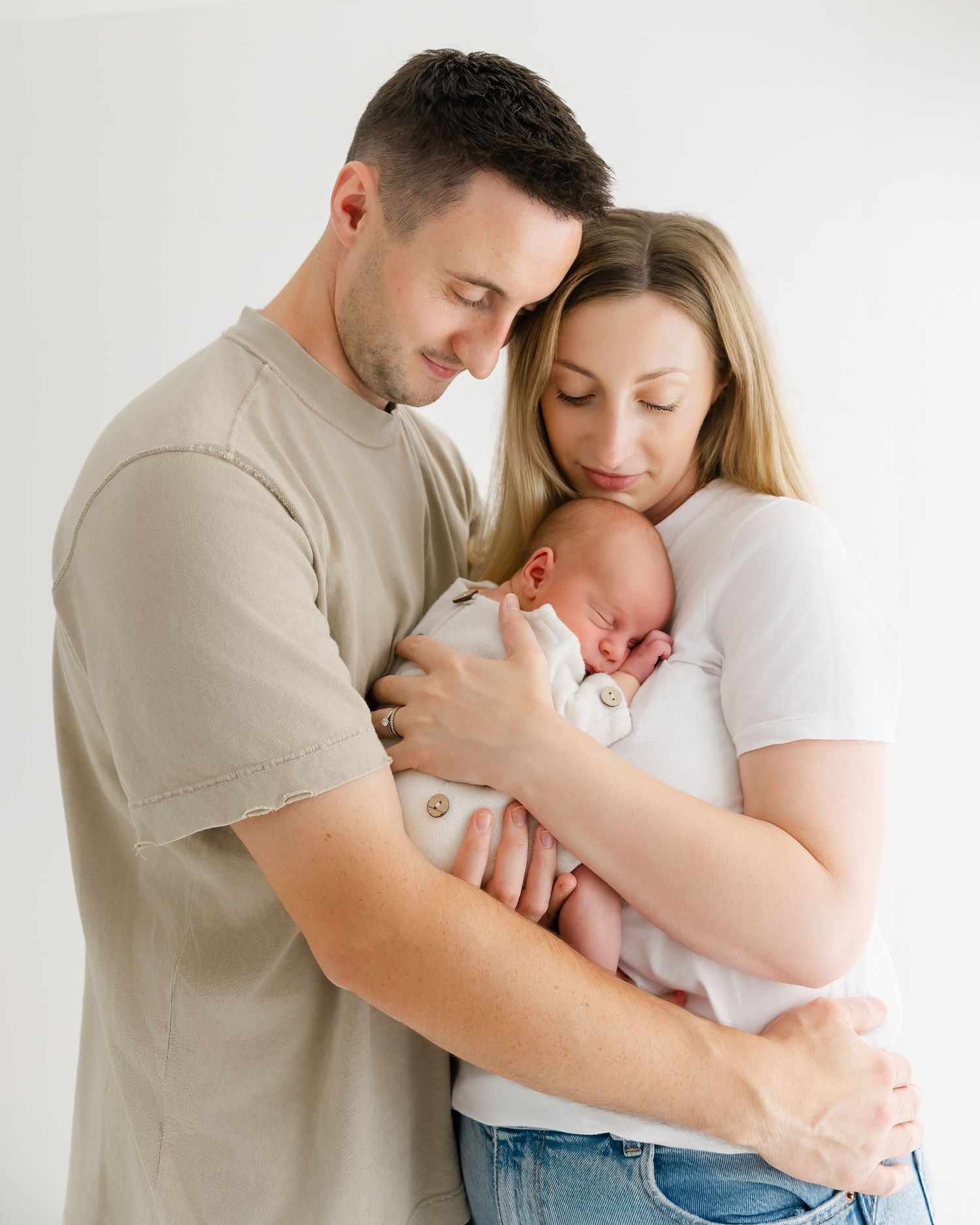 A couple tenderly holds their newborn baby together in a bright studio, wearing casual neutral tones.