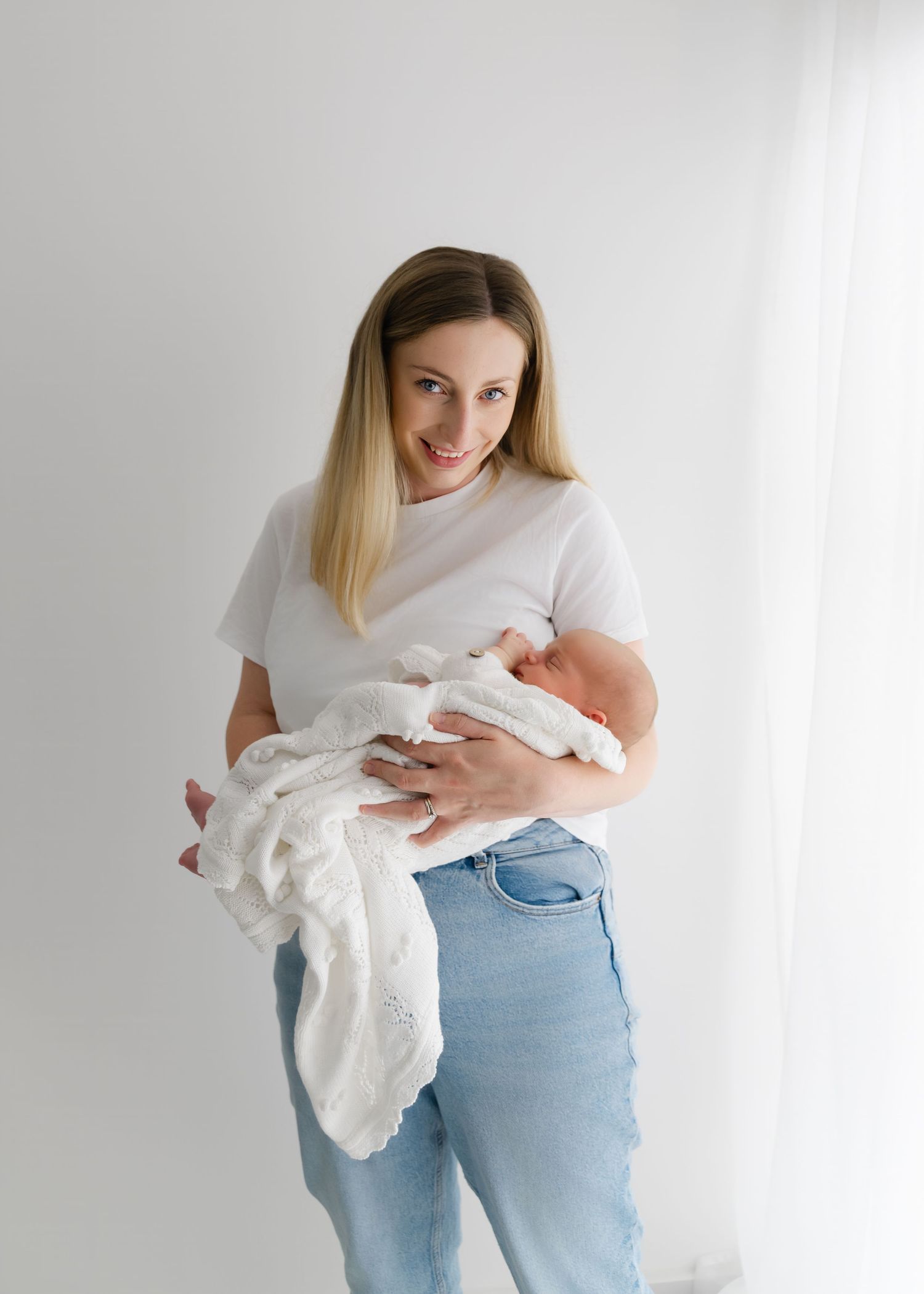 Smiling blonde woman in white t-shirt and jeans holding a sleeping newborn baby wrapped in white blanket.