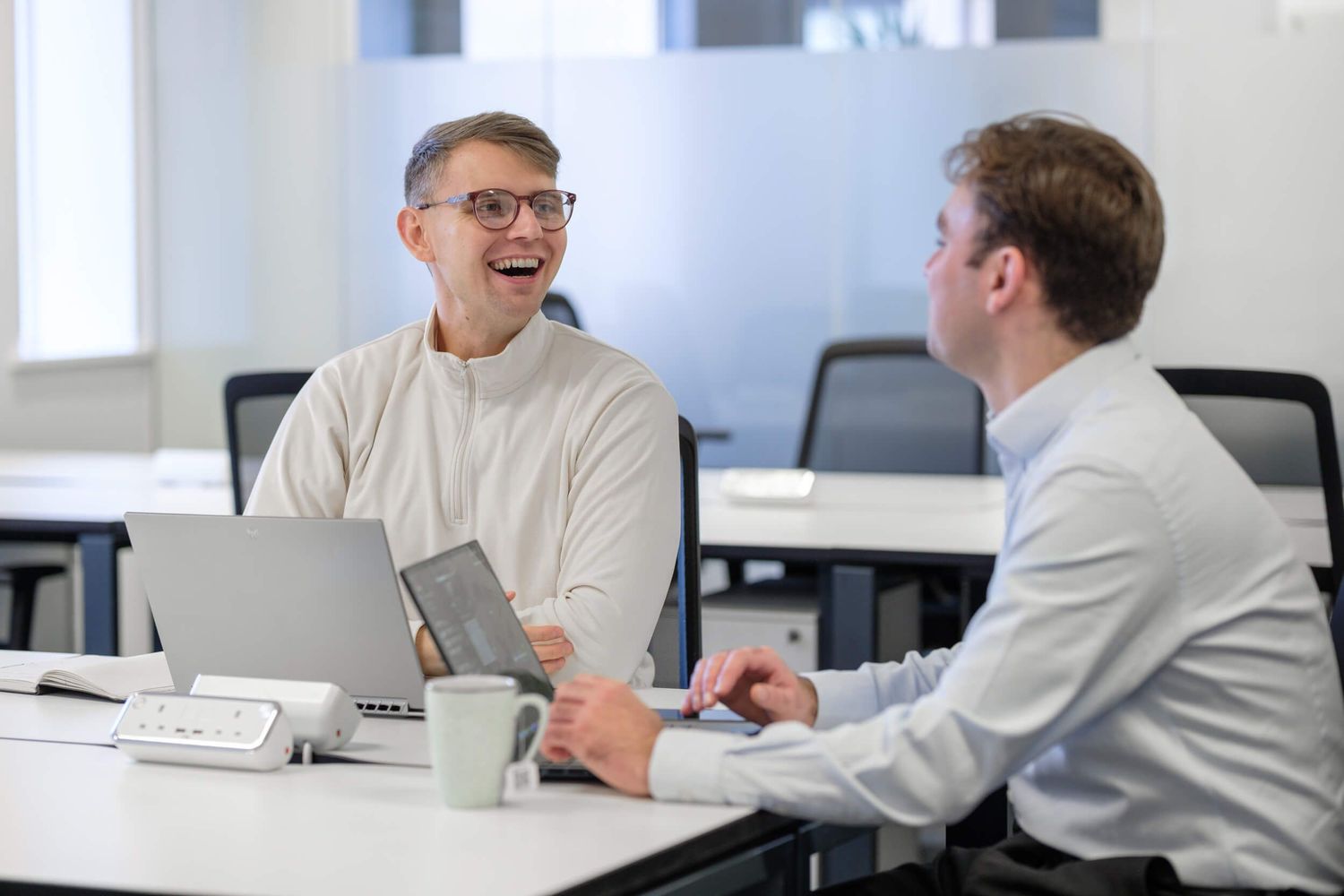 Two businessmen laughing and collaborating at a modern office desk with laptops and a tablet.