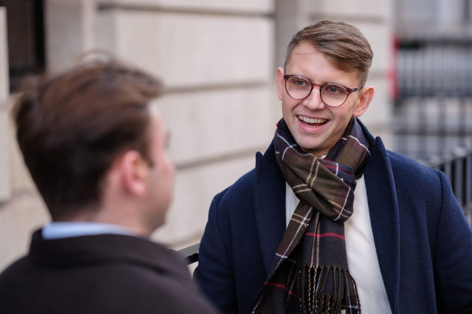 Two people in conversation outdoors, one wearing glasses and a plaid scarf with a navy coat smiling during discussion.