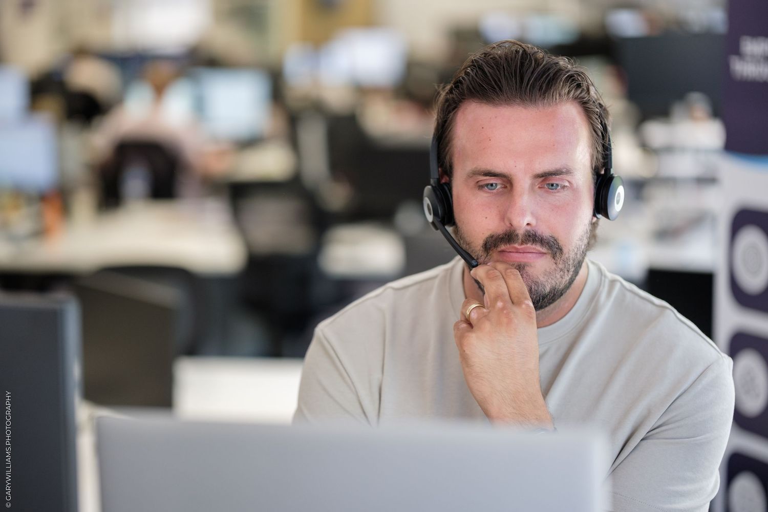 A focused male call center agent wearing a headset sits at his desk in a modern office environment.