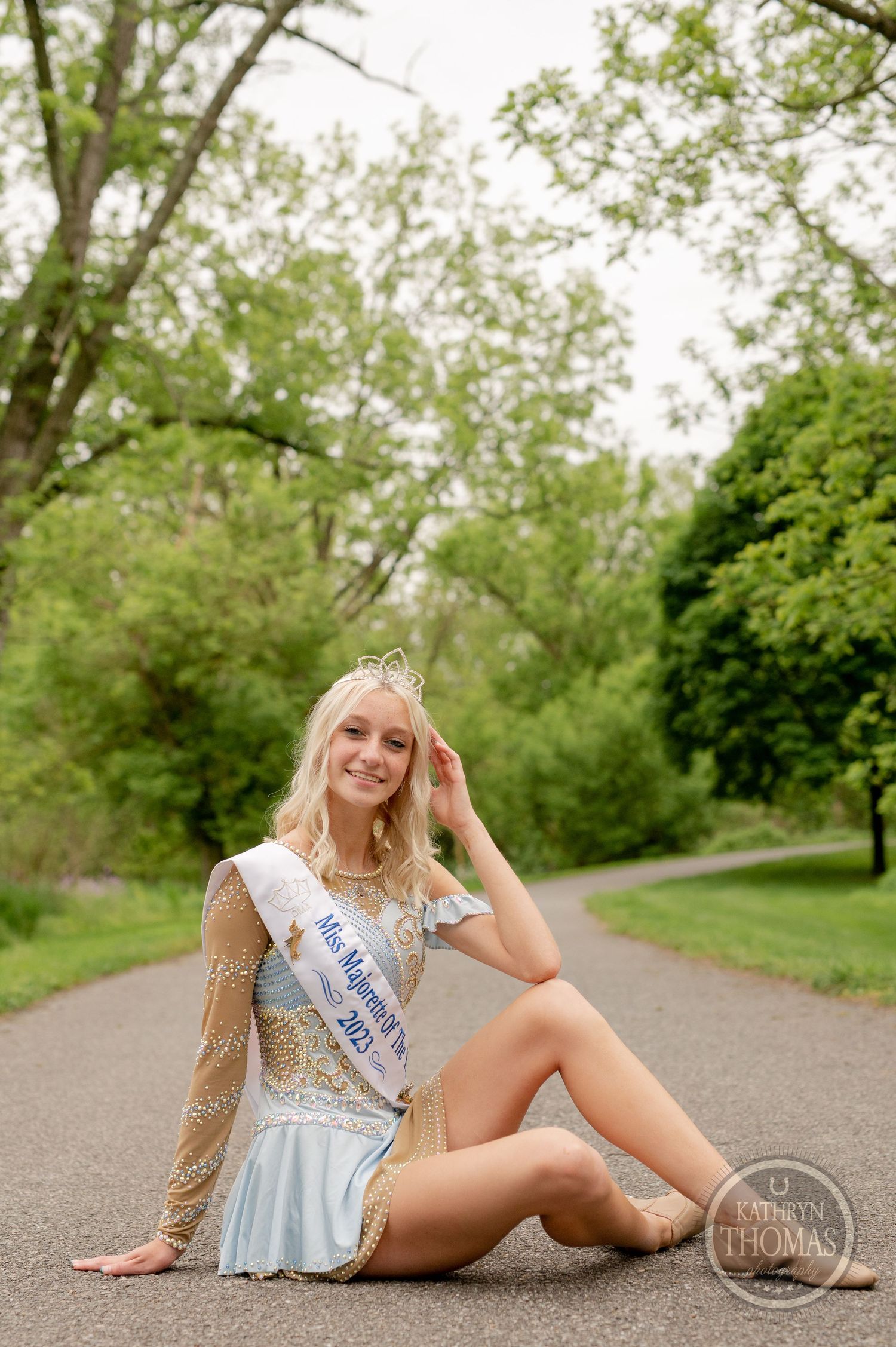 Pageant contestant in an elegant white dress sits gracefully on a garden path with a crown and sash.