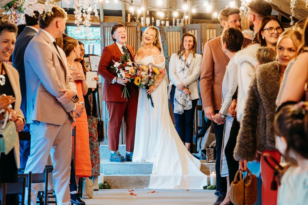 A wedding ceremony takes place in a rustic venue with warm lighting and guests gathered around a couple on wooden steps.