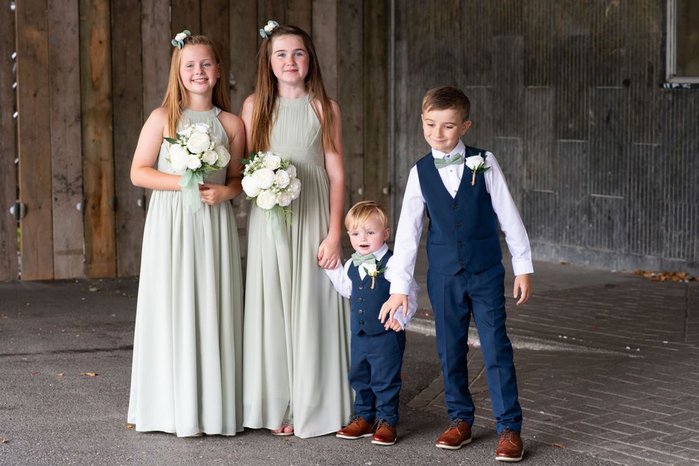 Wedding party members in sage green dresses pose with two young ring bearers at a rustic venue.