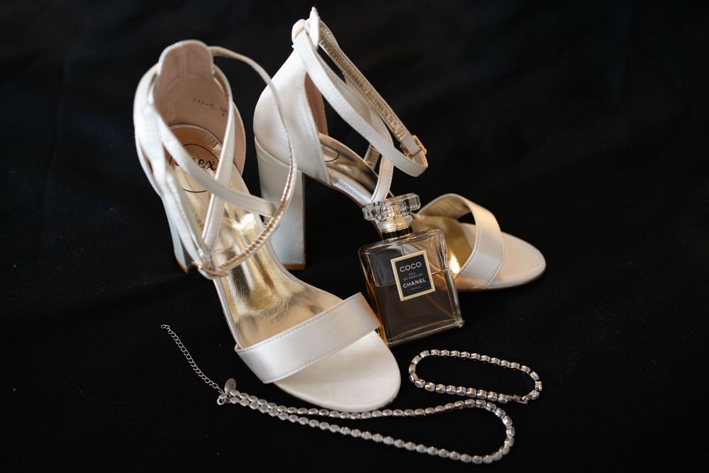 White bridal heels with ankle straps and delicate pearl necklace on dark background.