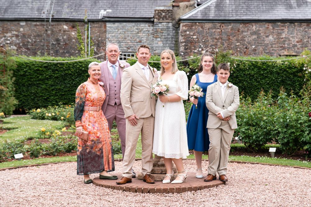 Small wedding party poses together on gravel path with stone walls and manicured hedges in garden setting.