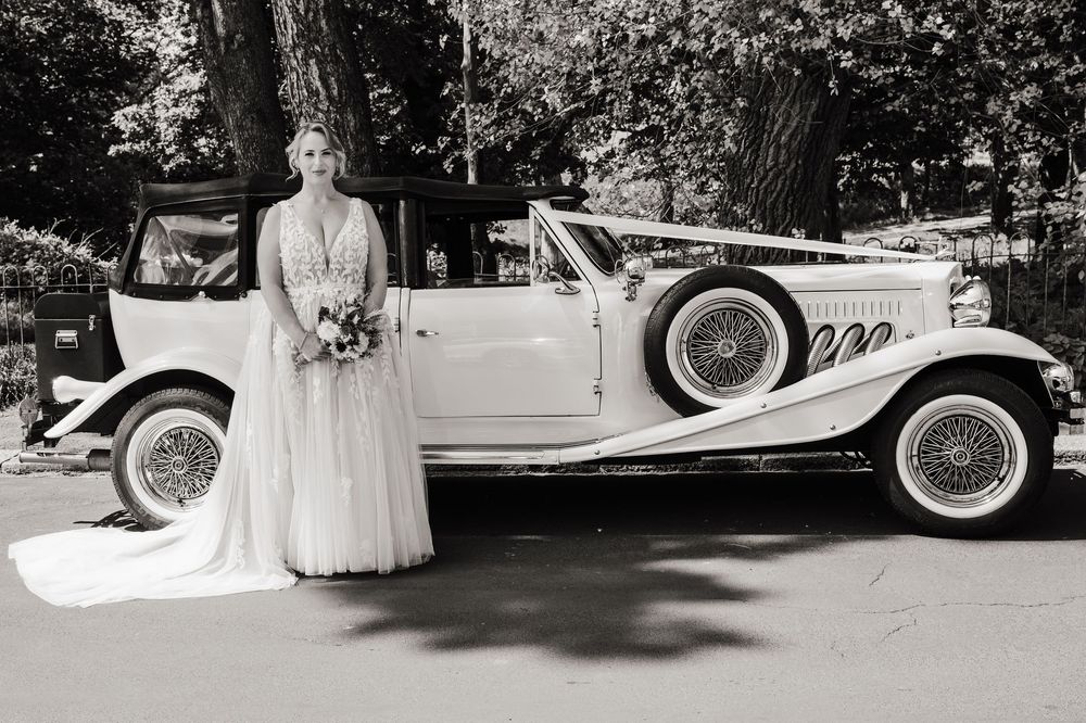 Bride in white wedding gown posing beside a vintage white convertible car, black and white photo.
