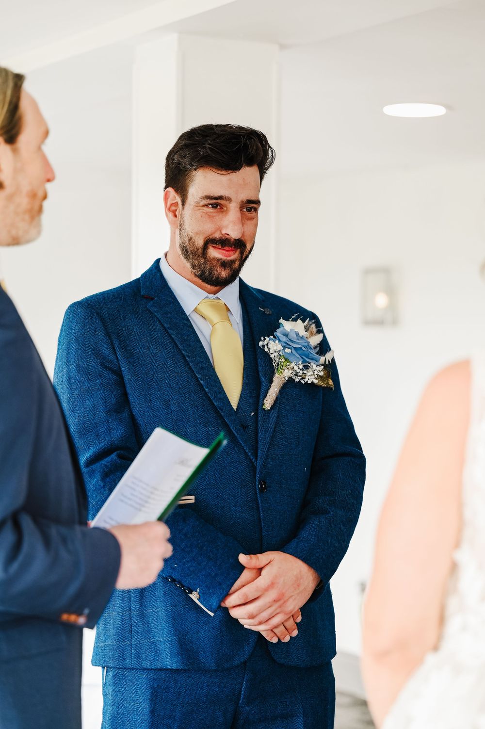 Groom in blue tweed suit with yellow tie and floral boutonniere smiling during wedding ceremony.