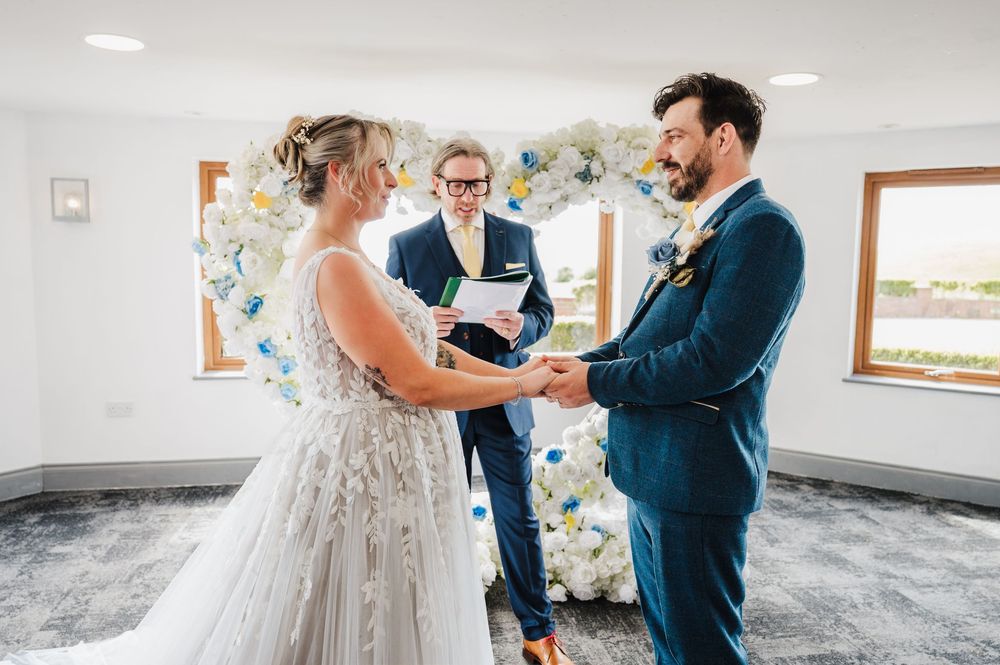 Couple exchanges vows at indoor wedding ceremony with floral arch backdrop and officiant present.