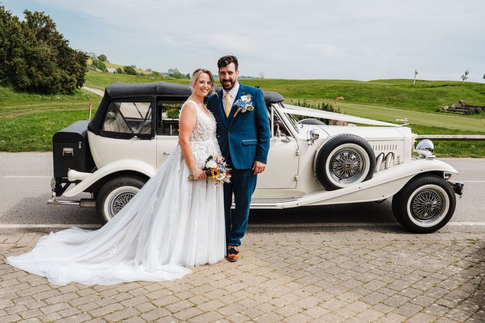 Bride and groom posing in front of a vintage white Beauford wedding car on a sunny day.