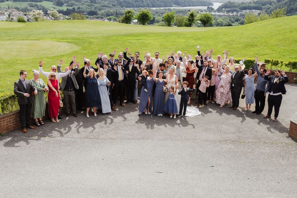 Large wedding party group photo outdoors with scenic green hills and water in the background.